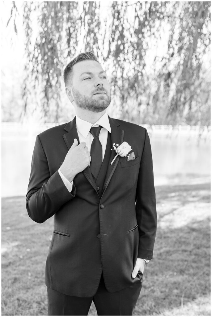 black and white photo of groom looking off into distance and holding edge of his suit coat at pond view farm in maryland