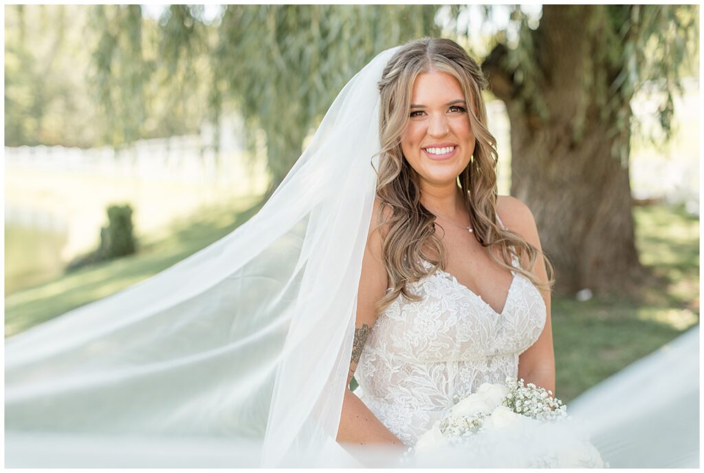 bride smiling at camera with veil extended behind her on sunny summer day outside at pond view farm