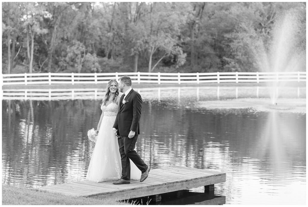 black and white photo of couple walking on wooden dock of pond and smiling at each other at pond view farm in maryland