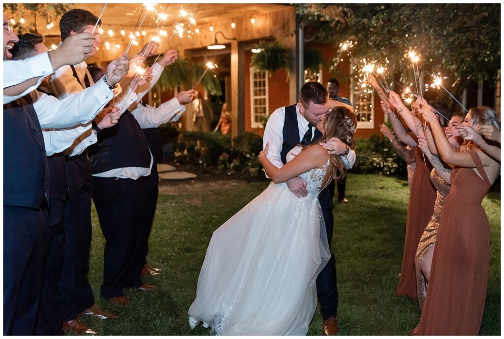 groom kissing his bride and dipping her back as they exit reception at nighttime with guests holding sparklers at pond view farm