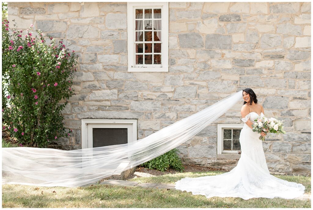 bride on right side of the photo looking back over her right shoulder with her very long veil floating behind her by stone building in lancaster pennsylvania