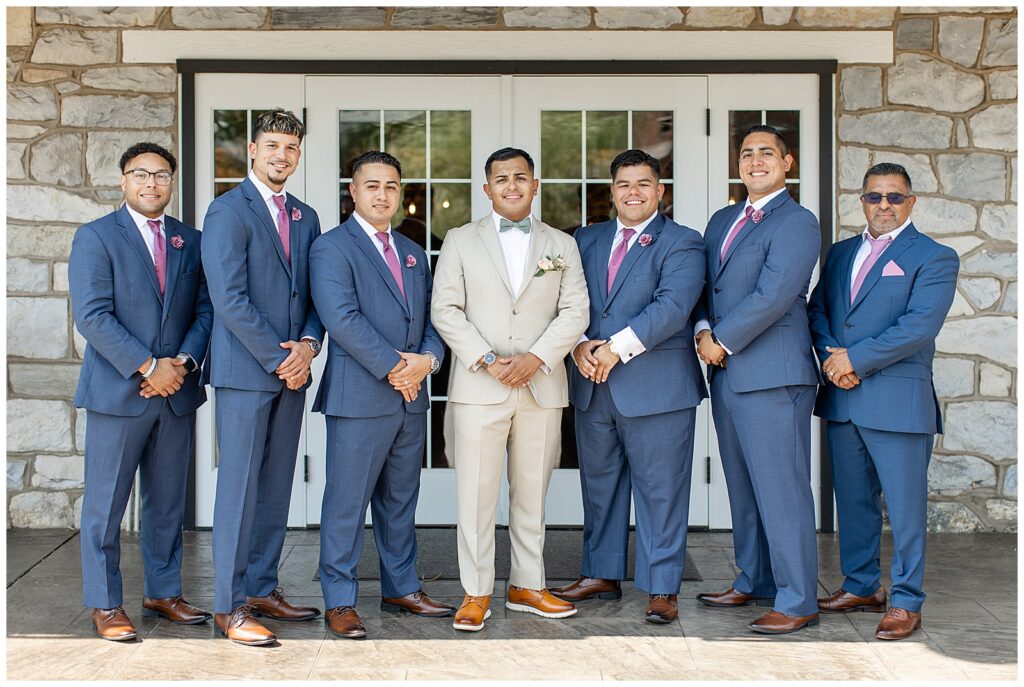 groom with his six groomsmen in navy blue suits standing by white doors at the barn at silverstone