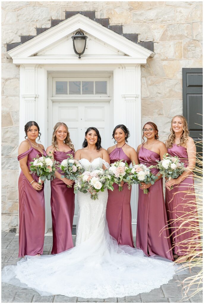 bride with her five bridesmaids in mauve gowns all holding bouquets and standing by white door of home at the barn at silverstone