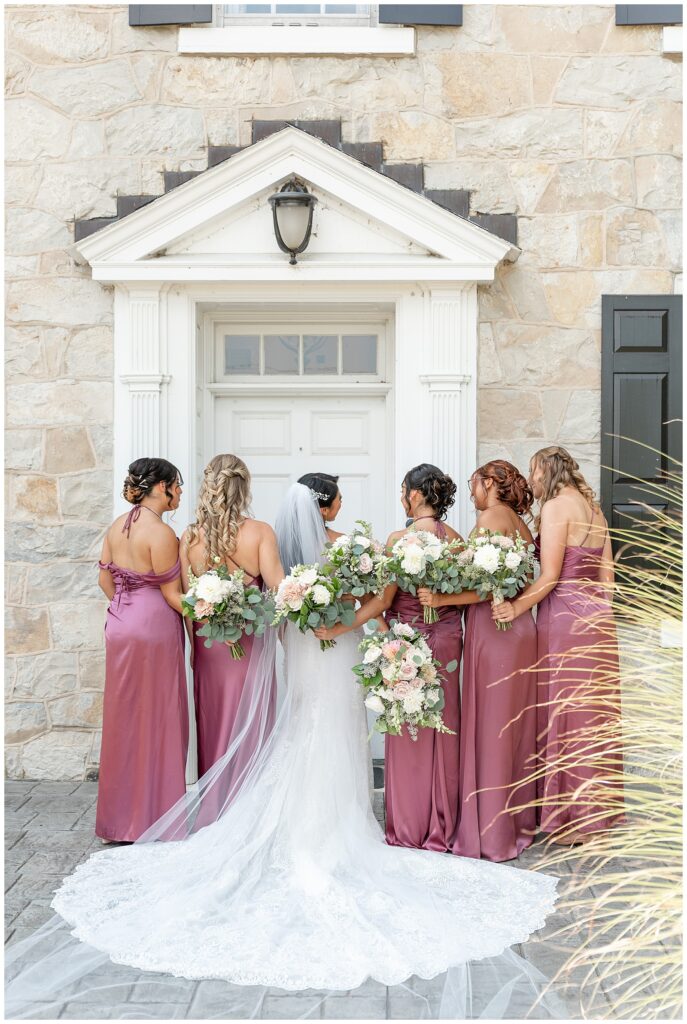 bride and bridesmaids with their backs toward the camera by white door of stone house at the barn at silverstone
