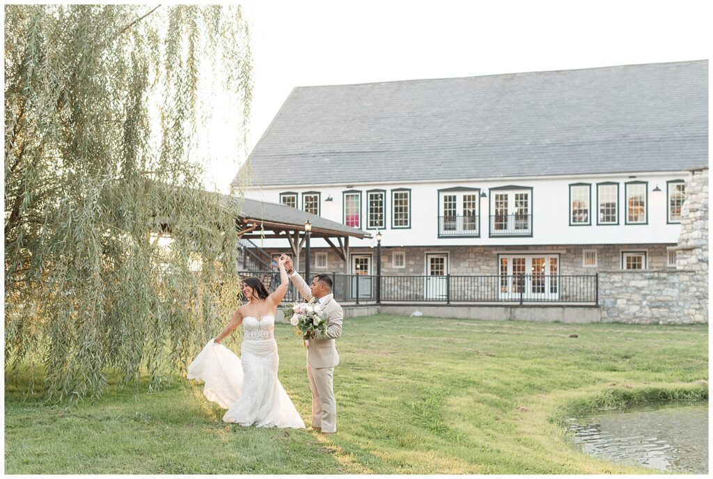groom twirling his bride under his right hand by willow tree in front of white barn at the barn at silverstone