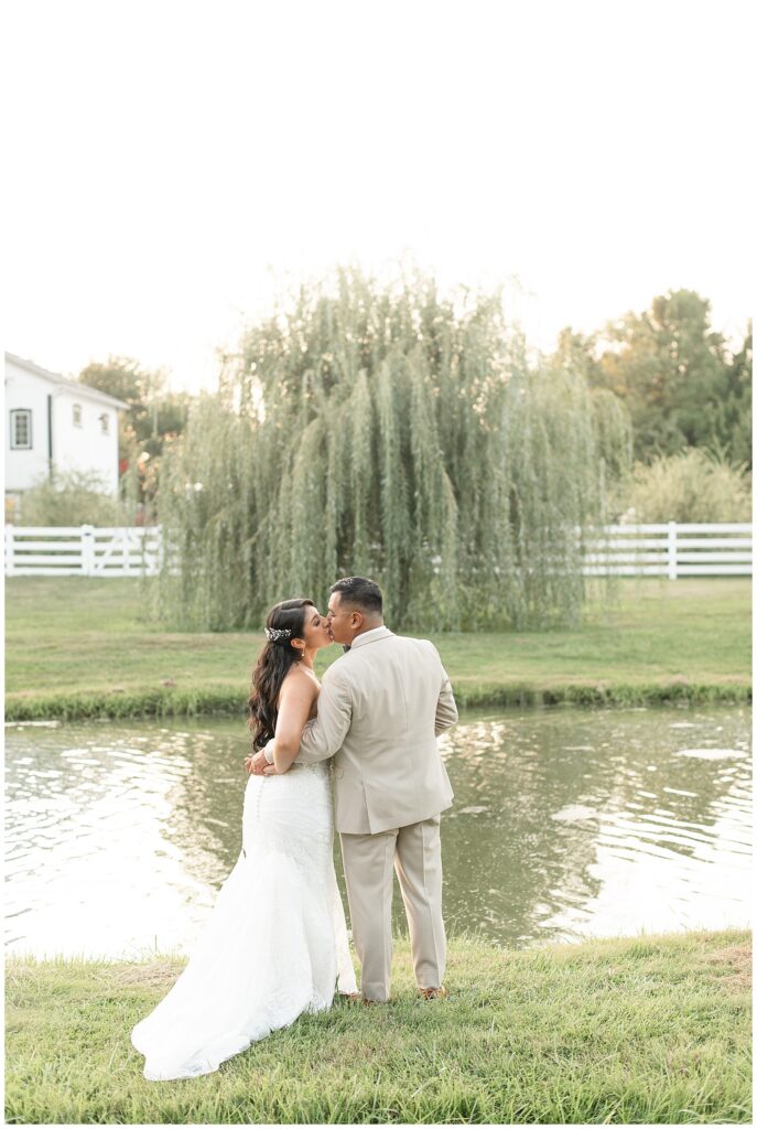 couple kissing with their backs toward camera by pond with willow tree in the background at the barn at silverstone