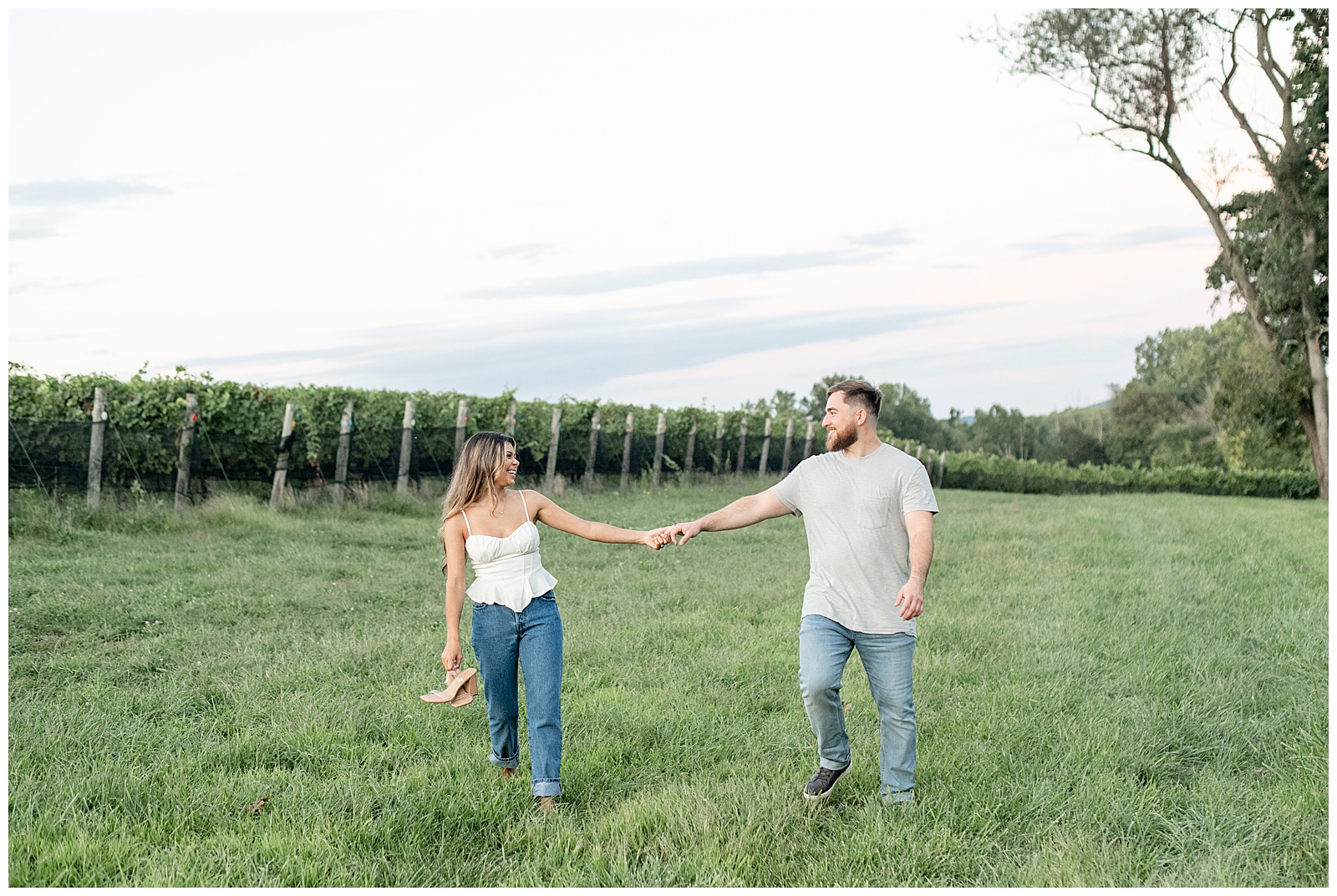 engaged couple holding extended hands as they walk in meadow and look at each other with vineyard in background at Domaine Pterion