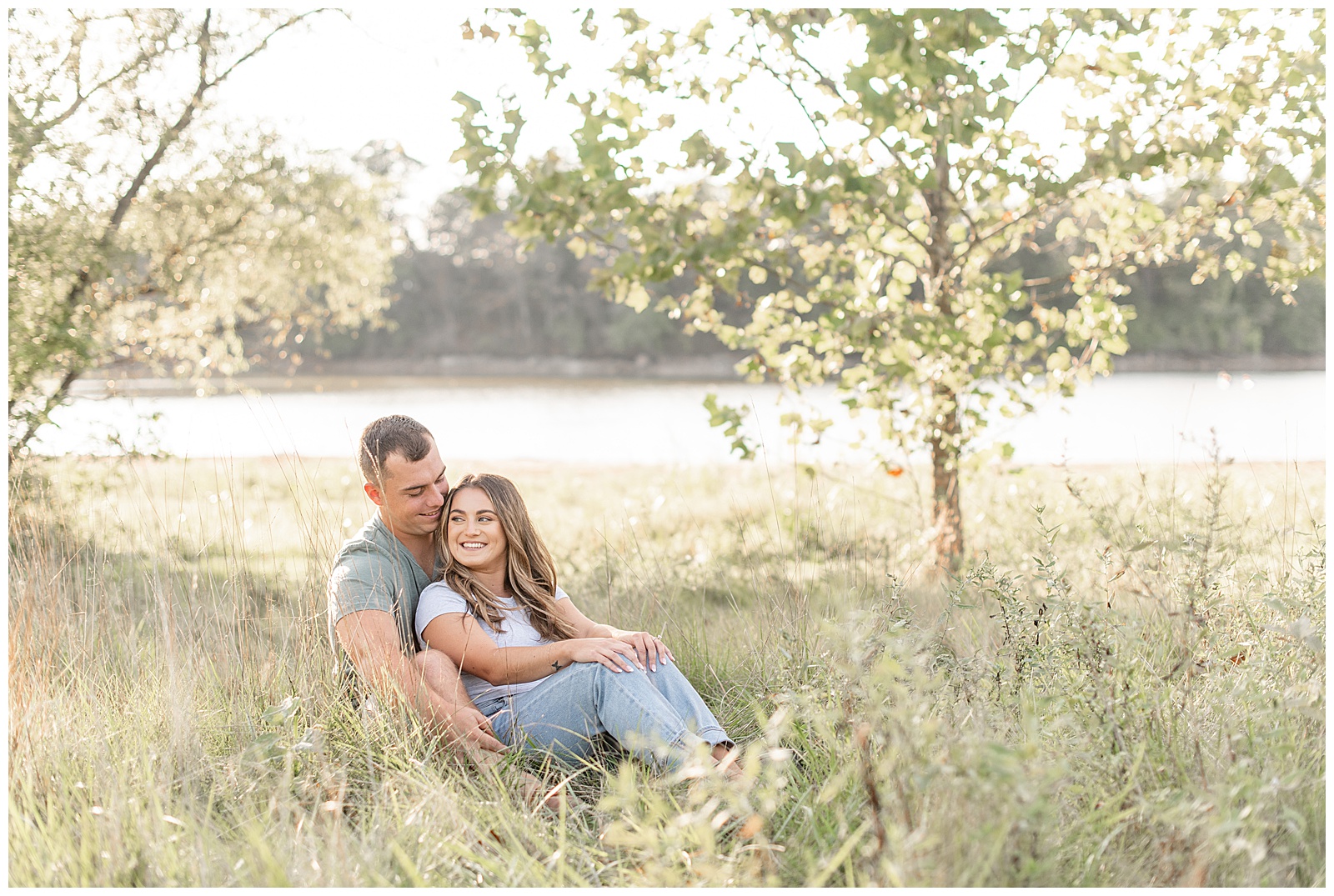 engaged couple sitting in tall wild grasses with woman leaning back against man at sunset with lake redman behind them