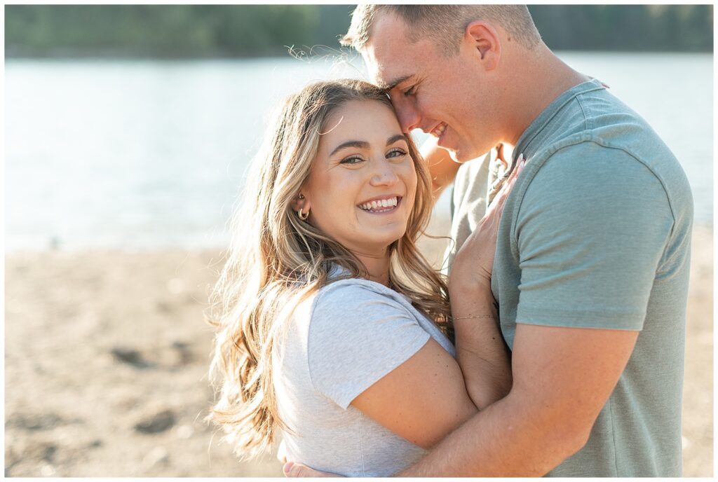 engaged couple hugging as girl smiles at camera and guy rests his forehead against her left temple on beach at lake redman