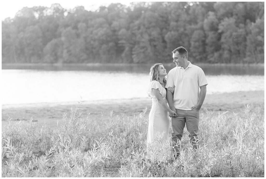 black and white photo of couple standing in wild grasses as woman looks up and smiles at man as she holds his right hand at lake redman