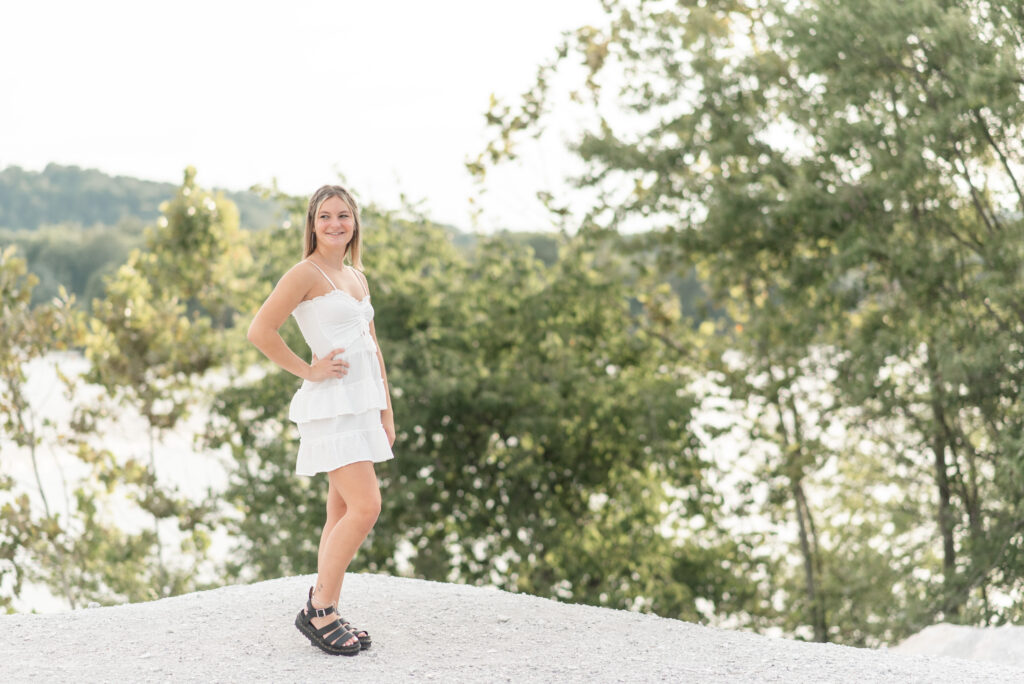senior girl in cute white dress with her right shoulder towards camera and right hand on hip standing on large rock at white cliffs of conoy