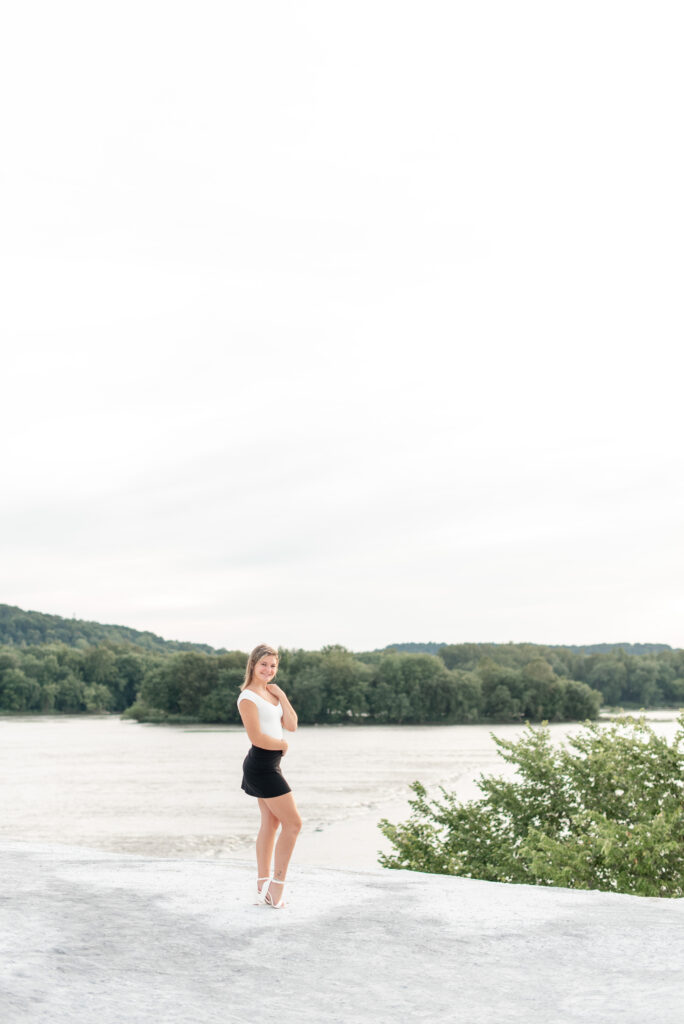 senior girl in white top and back mini skirt with right knee popped with susquehanna river in background at the white cliffs of conoy