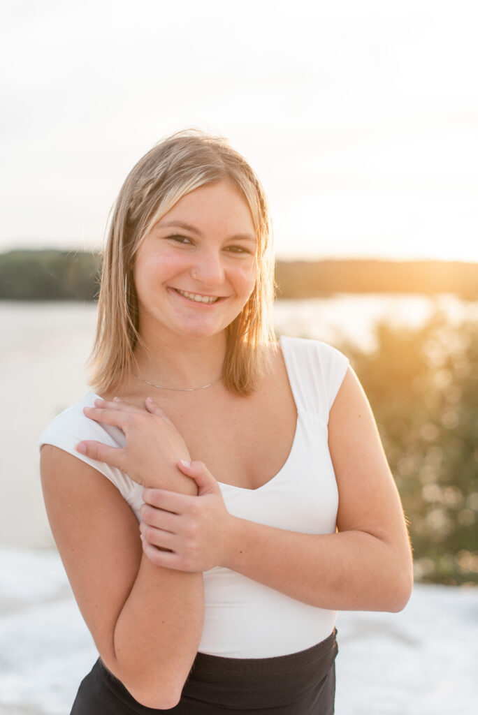 closeup of senior girl with right hand on right shoulder smiling at camera with the glow of sunset behind her at the white cliffs of conoy