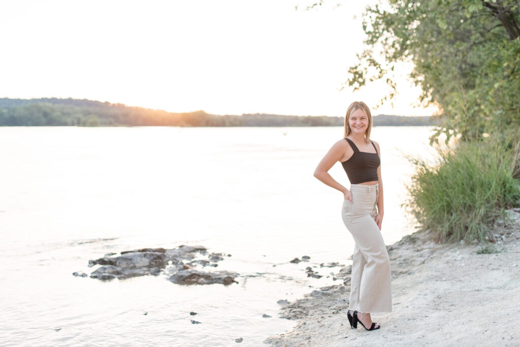 senior girl standing along the edge of susquehanna river in black tank top and khaki pants at sunset at the white cliffs of conoy