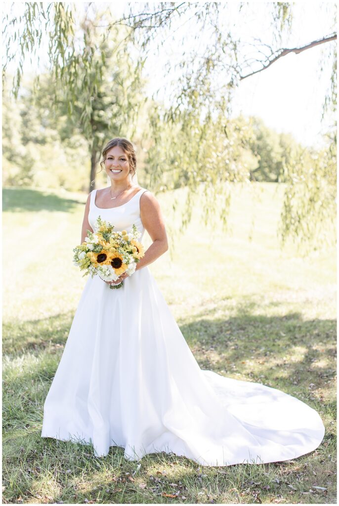 bride holding her bouquet of sunflowers with her dress train on display outside at brick gables
