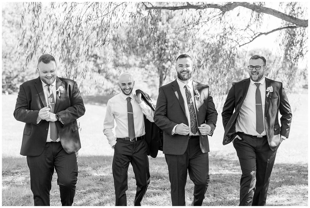 black and white photo of groom and his groomsmen walking towards camera and smiling outside at brick gables