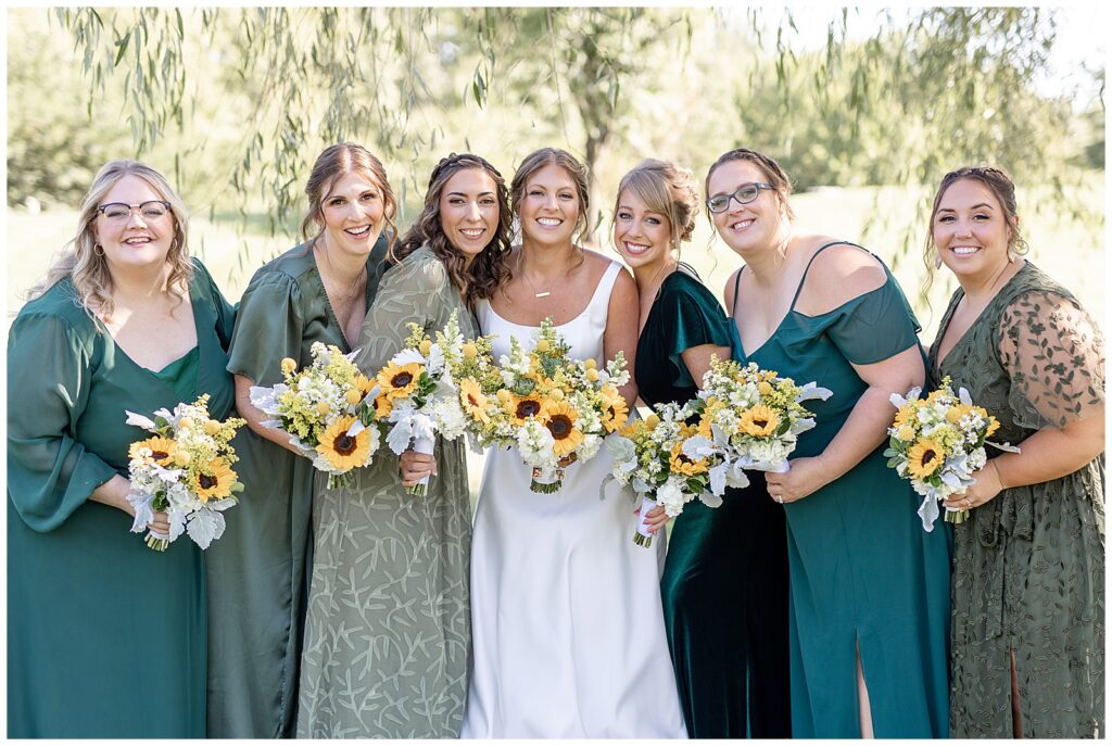 bride surrounded by her bridesmaids who are all wearing dresses in different shades of dark green and holding sunflower bouquets