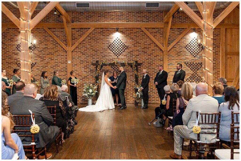 indoor wedding ceremony inside brick barn as couple holds hands and faces each other at brick gables