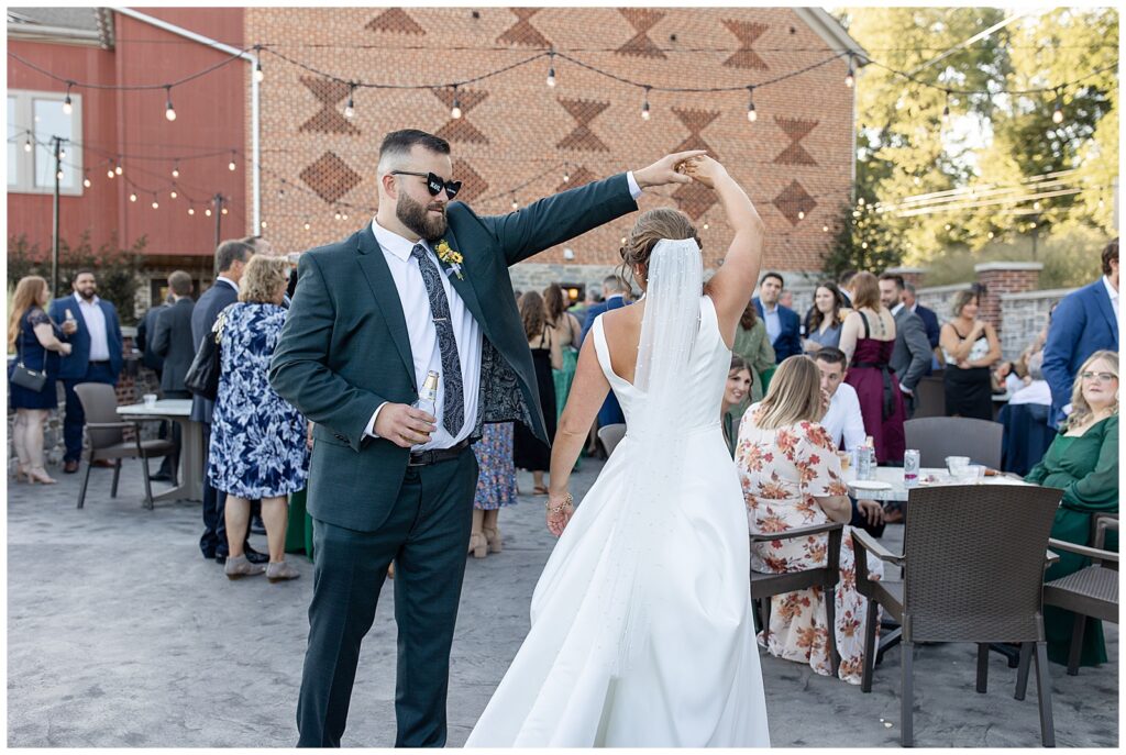 groom wearing sunglasses and twirling his bride outdoors with guests around them at brick gables