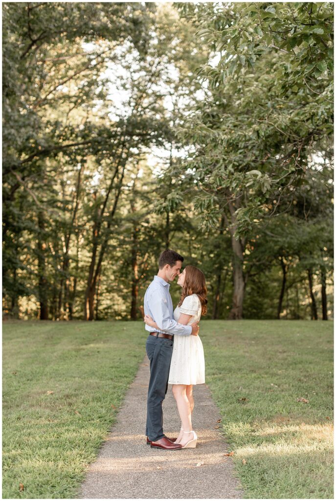 guy wearing light blue dress shirt and dark blue pants and girl wearing short white dress hugging tightly almost kissing on pathway at valley garden park
