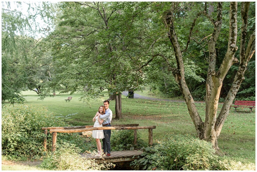 engaged couple standing on cute rustic wooden bridge with guy hugging girl from behind at valley garden park in delaware