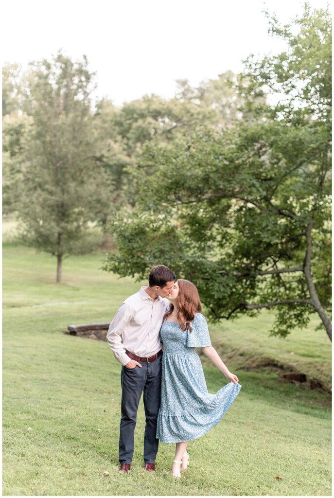 engage couple kissing as girl holds the edge of her long blue and white floral dress with her left hand at valley garden park