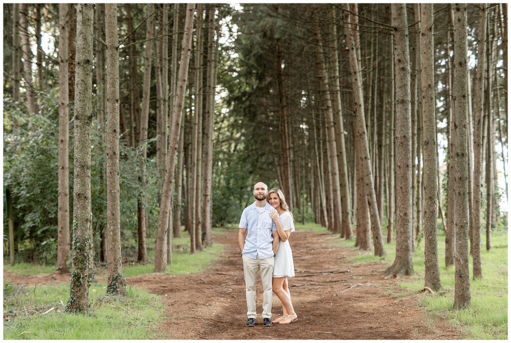 engaged couple standing close together on pathway in between rows of evergreen trees at overlook park