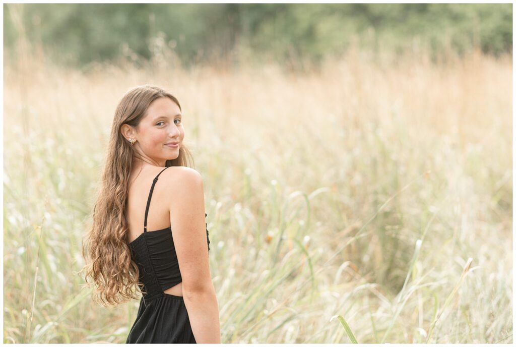 senior girl with right shoulder towards camera smiling wearing black romper in tall wild grasses at hibernia park