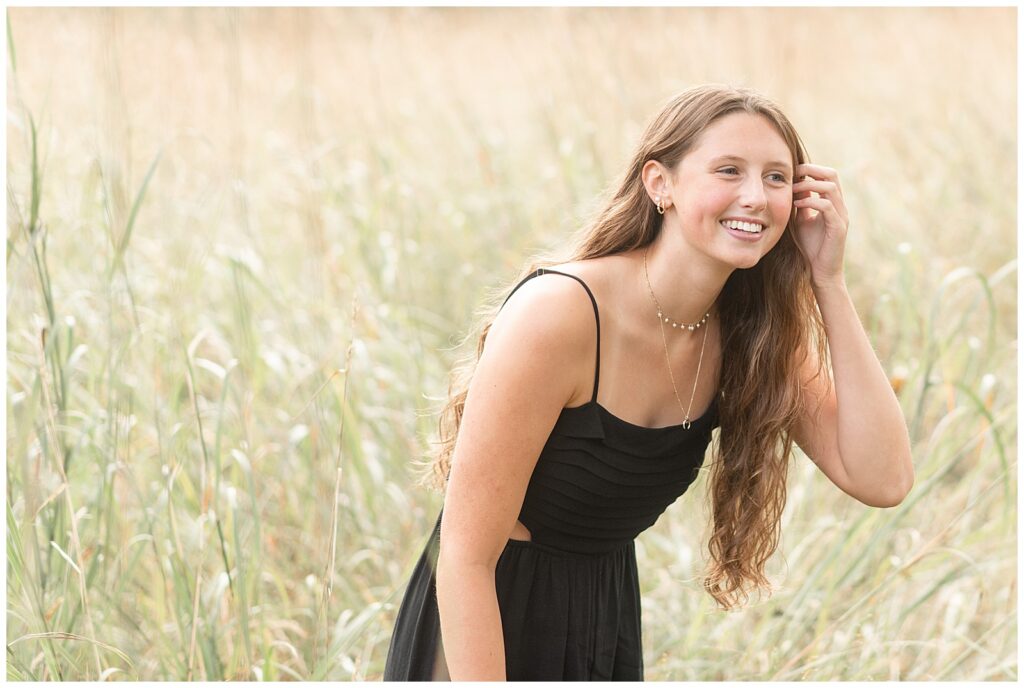 senior girl leaning forward tucking hair behind her left ear and smiling but not looking at camera in tall grasses at hibernia park