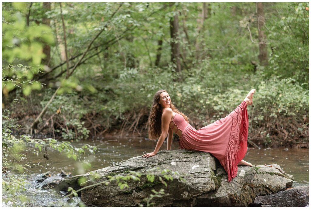 senior girl in long dark pink dress sitting on large rock and extending right leg into the air by stream at hibernia park
