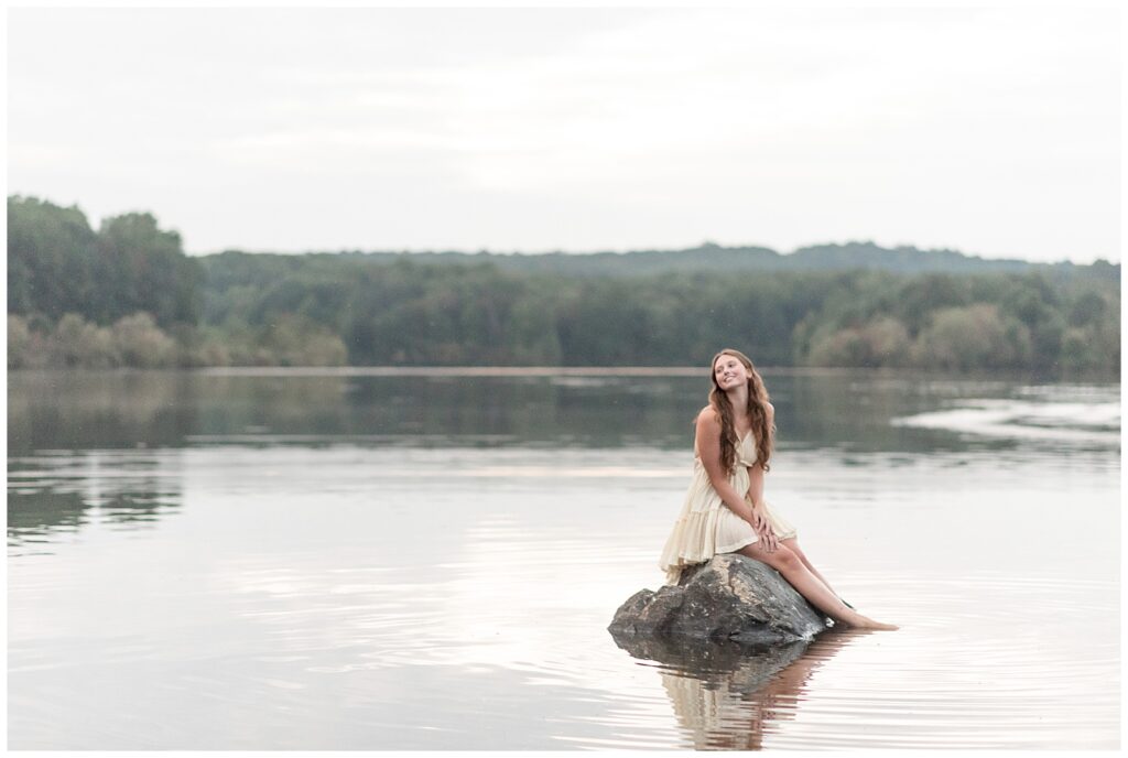 senior girl in ivory dress sitting on large rock in the middle of lake at hibernia park on cloudy fall evening