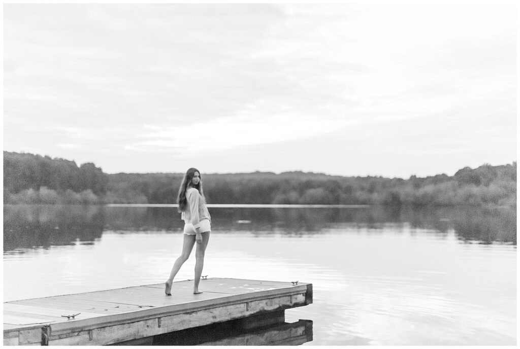 black and white photo of senior girl walking towards end of dock and looking back at camera by lake at hibernia park in pennsylvania