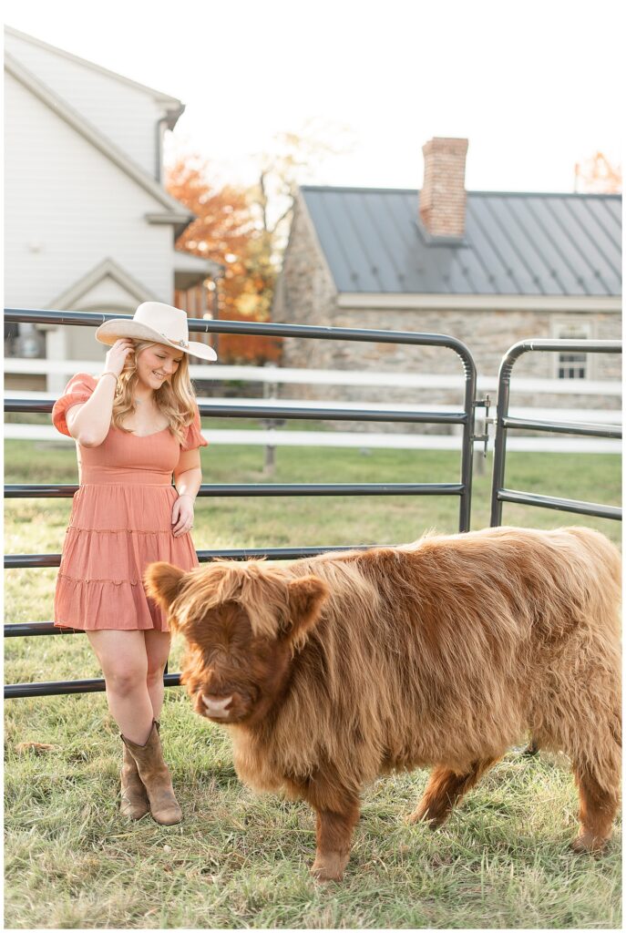 senior girl in rust colored dress and white cowboy hat by highland cow in york pennsylvania