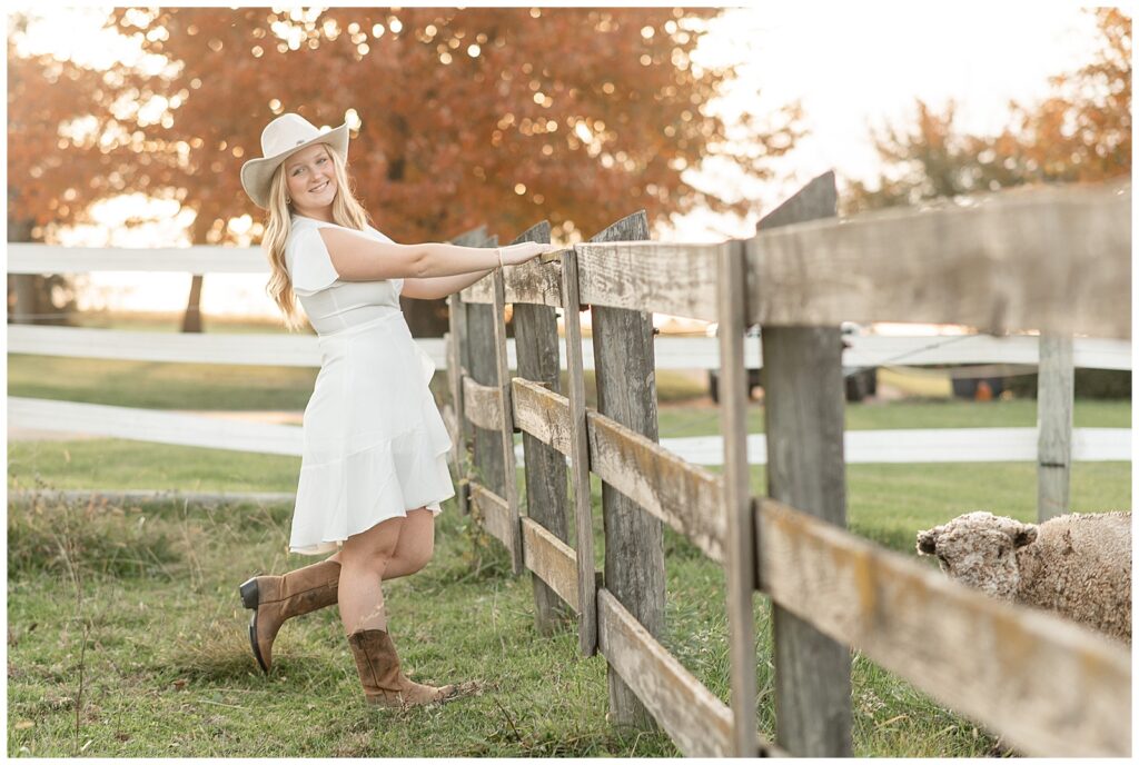 senior girl in white dress and white cowboy hat facing and holding onto wooden fence with both hands at fall sunset in york pennsylvania
