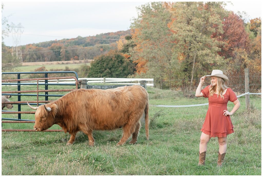 senior girl holding brim of her white cowgirl hat walking alongside large highland cow in pasture in york pa