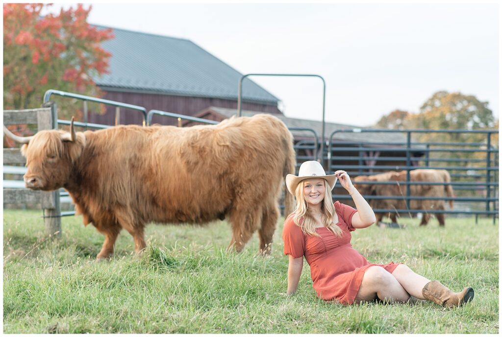 senior girl sitting in grass with highland cow behind her holding brim of at at farm in york pennsylvania