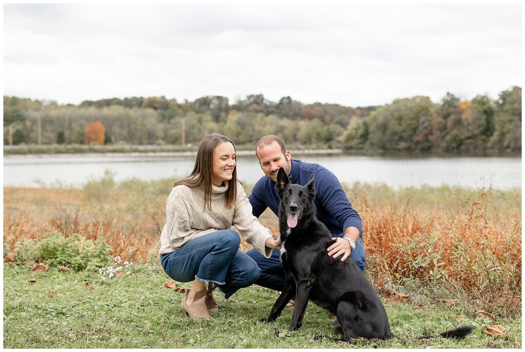 engaged couple crouched down petting their large black dog by lake redman in york pennsylania