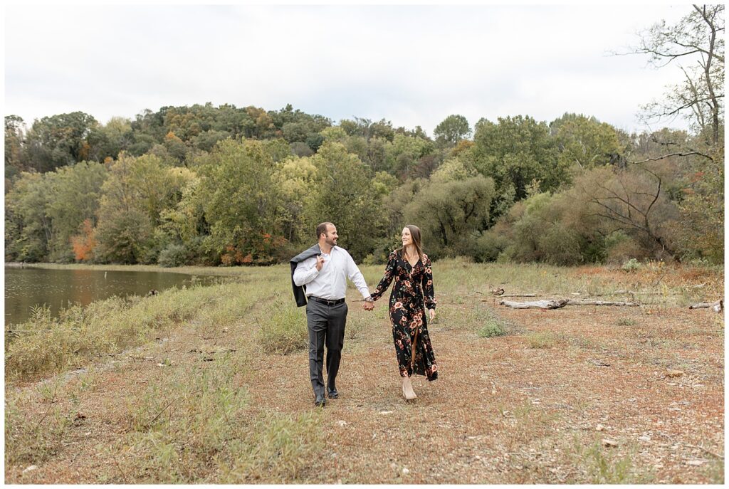 engaged couple holding hands and walking towards camera with guy's suit coat slung over his right shoulder at lake redman