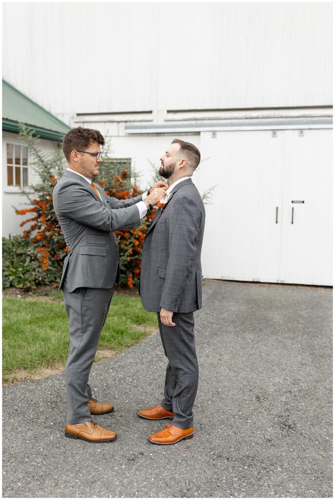 best man adjusts groom's tie and they're both wearing dark gray suits and standing by white barn at lakefield weddings