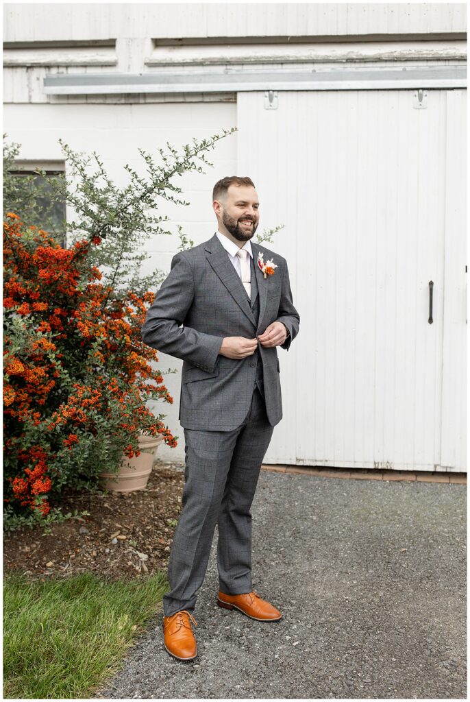 groom looking off into distance and smiling as he button his dark gray suit coat by bush with orange berries at lakefield weddings