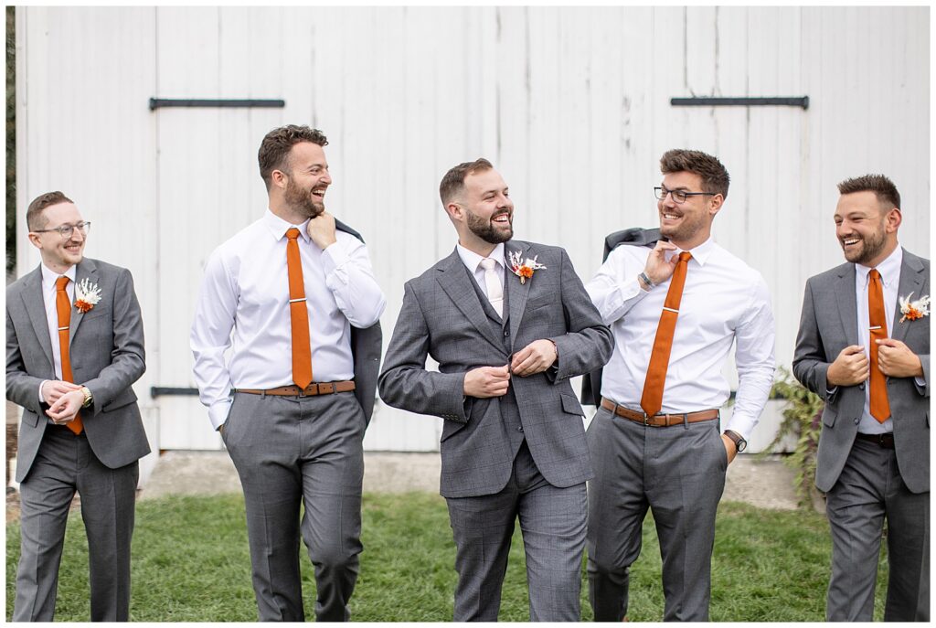 groom is buttoning his suit and looking to the left smiling as he walks towards camera with a groomsmen on either side of him in manheim pennsylvania