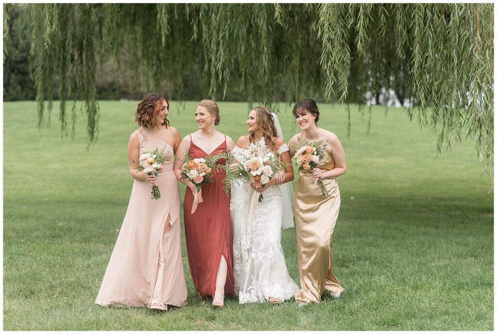bride walking towards camera in white lace gown with her three bridesmaids in shades of pink, gold, and rust dresses at lakefield weddings