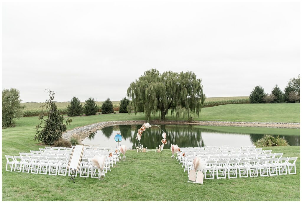 rows of white chairs facing lake with beautiful floral archway and willow tree behind lake on fall day at lakefield weddings