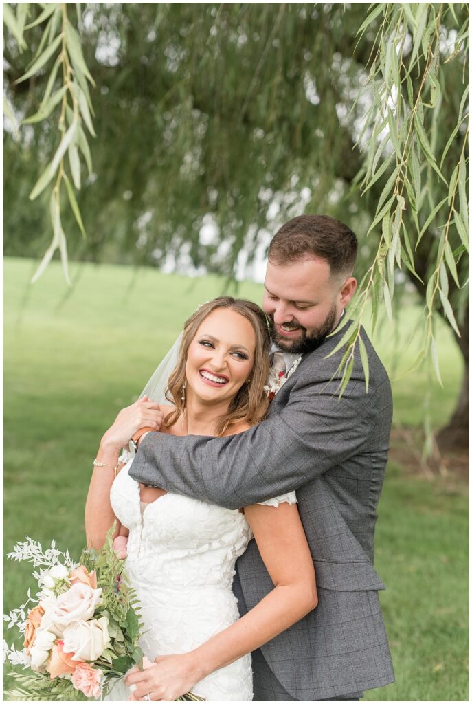 groom hugging his bride from behind and smiling as he looks down at her by willow tree at lakefield weddings