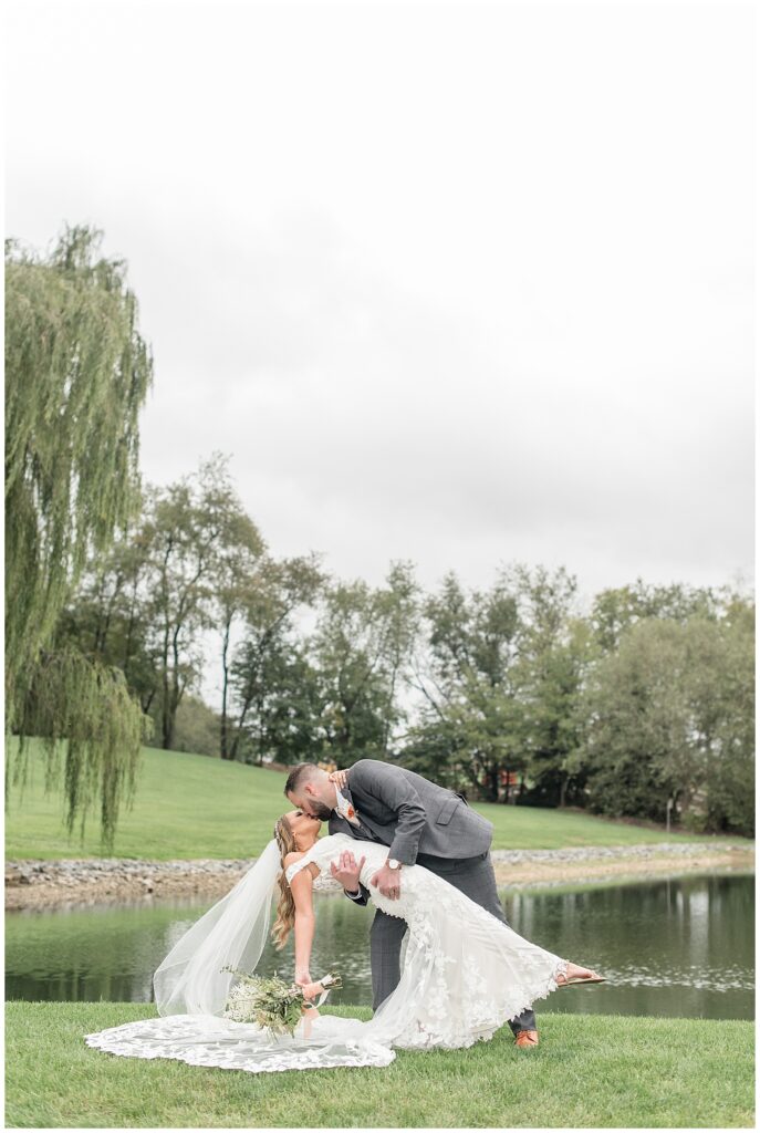 groom dipping back his bride and kissing her by pond and willow tree at lakefield weddings in manheim pa