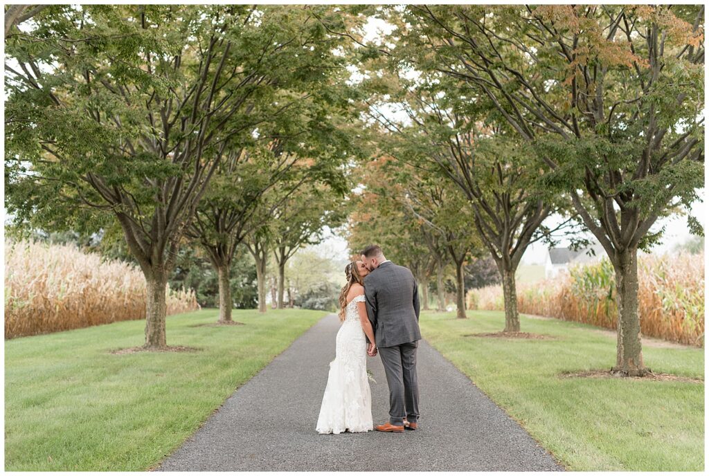 couple standing in middle of paved tree-lined pathway with backs toward camera and kissing at lakefield weddings