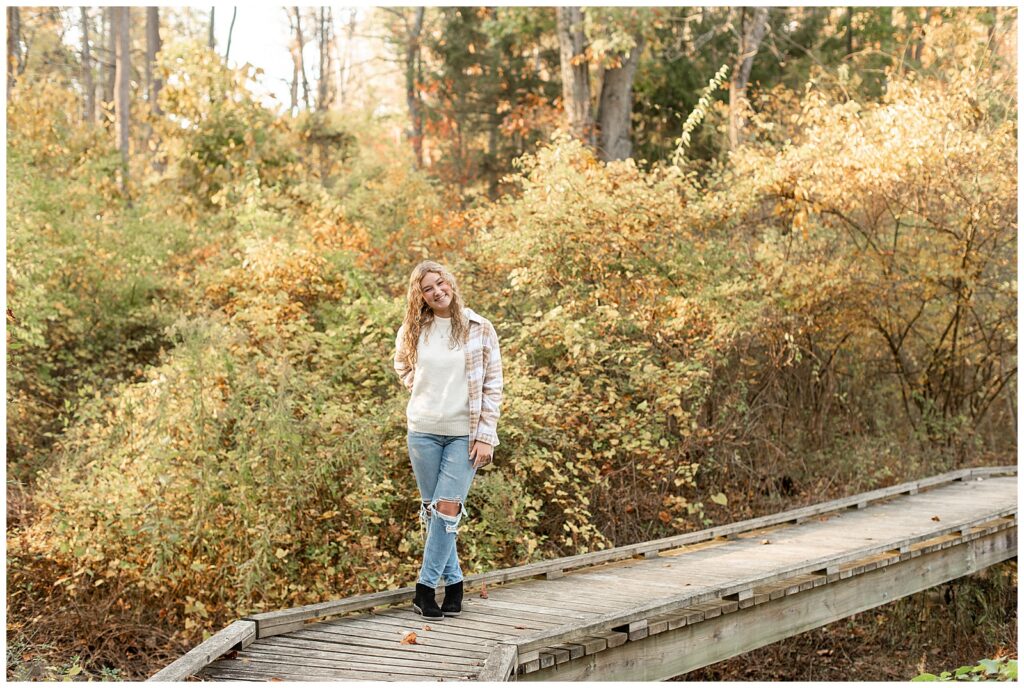 senior girl in oversized white and tan flannel shirt with blue jeans standing on wooden walkway at nolde forest