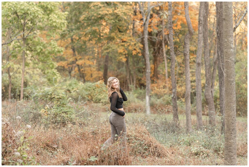 senior girl with back slightly toward camera looking back over right shoulder and smiling by colorful trees at nolde forest in reading pennsylvania