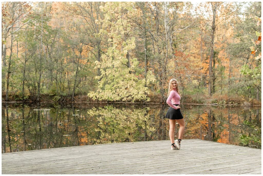 senior girl in pink top with short black skirt standing on wooden dock by water and colorful trees at park in berks county pennsylvania