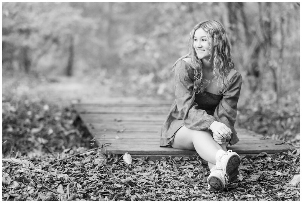 black and white photo of senior girl sitting on end of wooden pathway with legs extended out and looking right and smiling at nolde forest